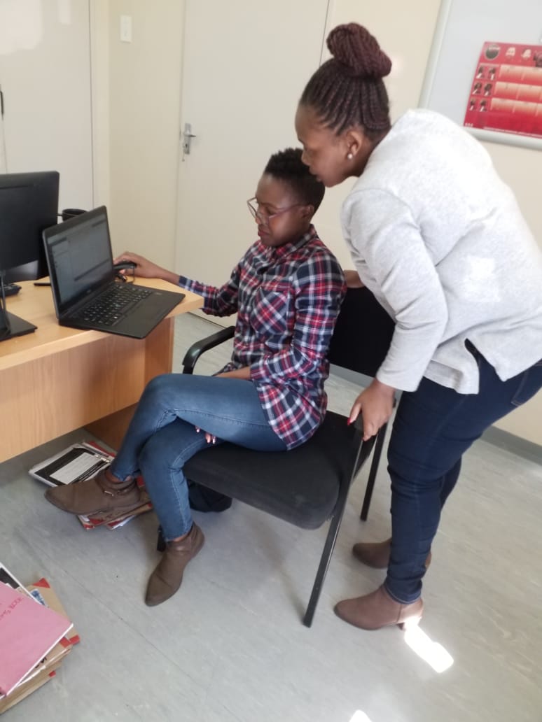 A member of staff is working an a laptop, and another is stood behind her looking at the screen