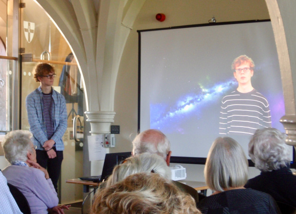 Joe Double stood in front of a seated audience, and alongside him is a video screen on which he is displayed