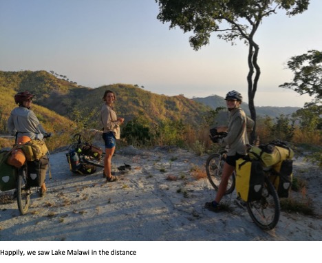 Three cyclists, stood next to their bikes, looking from an elevated location over hills with a lake just visible in the distance