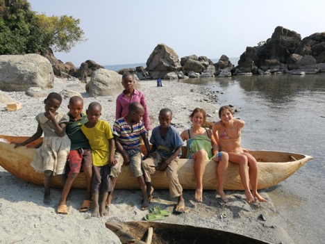 Two of the cyclists, sat on a wooden canoe on the edge of the lake with six local children