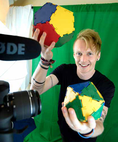 Tom rocks maths Tm Crawford holding multi-coloured three dimensional shapes in front of a camera, with a green-screen behind him