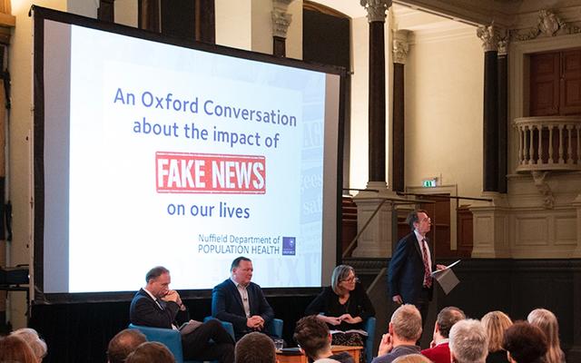 A view from within the audience of a stage with four speakers, and on the screen is a slide saying 'An Oxford Conversation about the impact of Fake News on our lives'