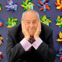 Gyles Brandreth, with his chin on his hands, in front of a wall of colourful windmills