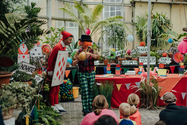 Children watch a colourful Alice in Wonderland–themed performance inside a greenhouse filled with plants and decorations