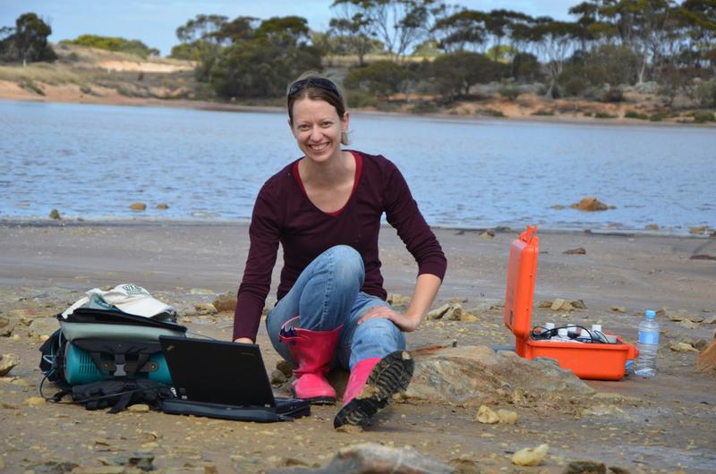Bethany Elhmann with a laptop and equipment, working on the beach of a lake