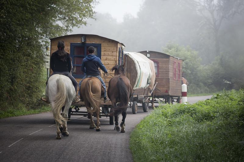 Three horses, two of which are being ridden, walking on a road behind three linked caravans, two of which are wooden and one is canvas