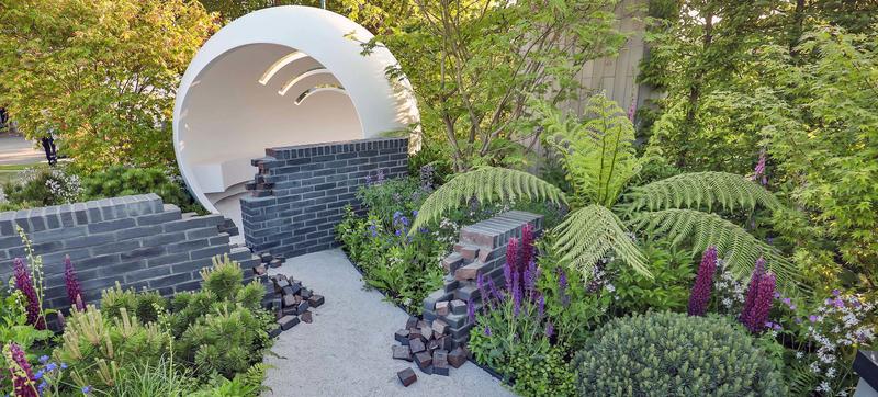 A garden installation, with a hollowed white sphere behind a broken grey brick wall, with a path leading to it, within green vegetation
