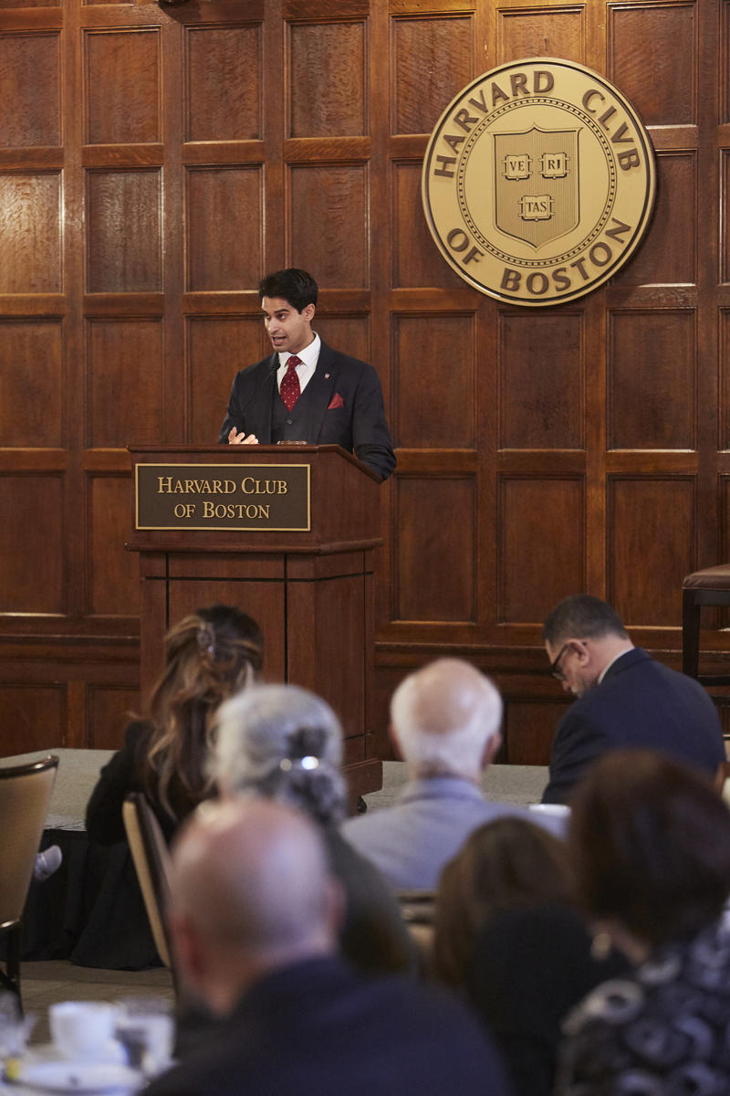 Jason Arora speaking behind a wooden lecturn with the Harvard Club of Boston seal behind him, and that wording on the lecturn