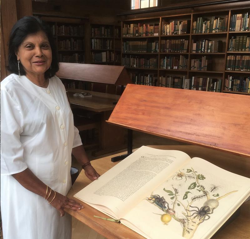 Jeyaraney Kathirithamby stood in a library next to a large open book, Merian’s Metamorphosis Insectorum Surinamensium