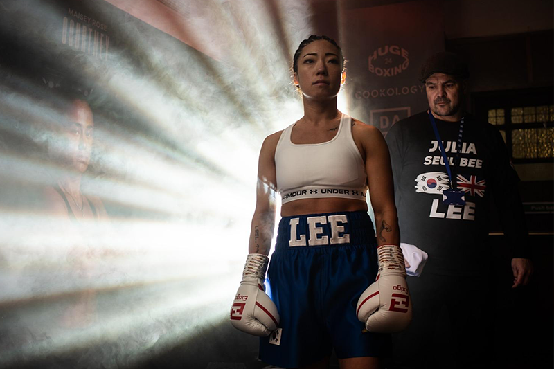 Julia Lee stands in a boxing ring wearing gloves and blue shorts, illuminated by dramatic light beams behind her as she prepares to compete, with a coach standing nearby