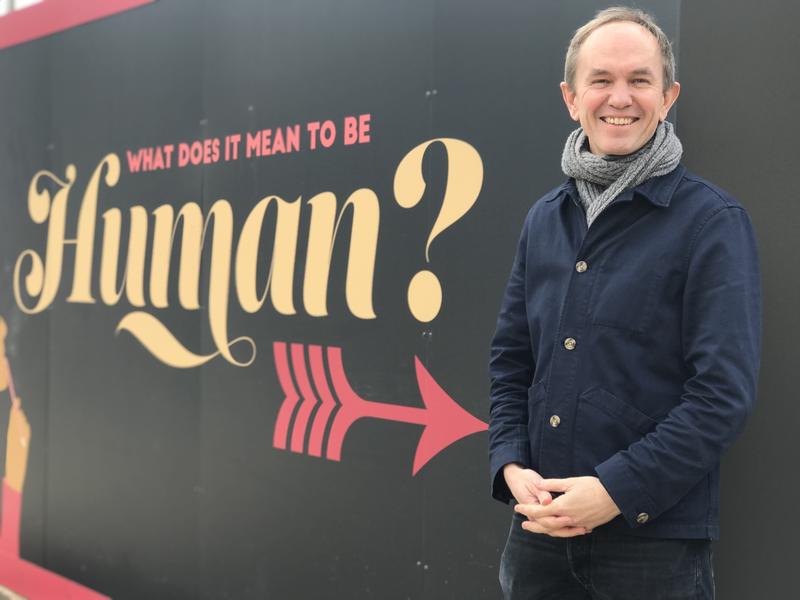 John Fulljames standing next to a hoarding with the words, 'What is it to be human'