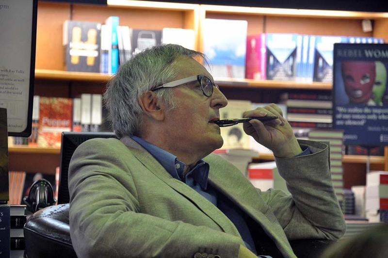 Professor John Gray, seated, listening to someone with a pen to his mouth