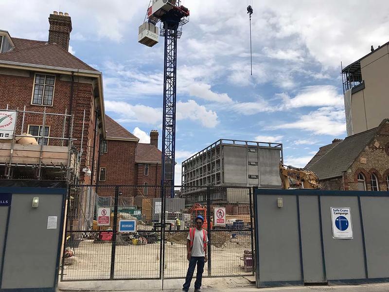 A workman stood outside gates to a empty construction site, in which a crane is situated