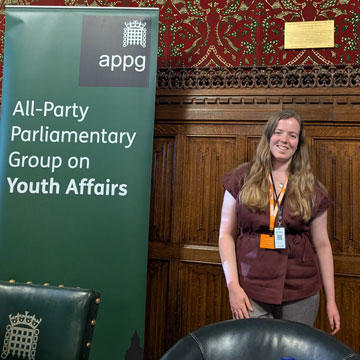 Carys Hoggan standing in a wood panelled room next to a banner for the all-party parliamentary group meeting on youth affairs