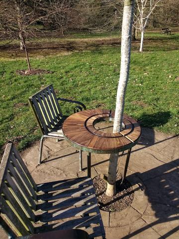 A picture of Havel's Place - a table surrounding a tree, with two chair, on a paved area 